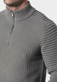 Grau gestrickter Pullover mit hohem Kragen, ausgestattet mit einem halben Reißverschluss. Texturierter Körper mit gerippten Schulterakzenten und einer taillierten Silhouette.