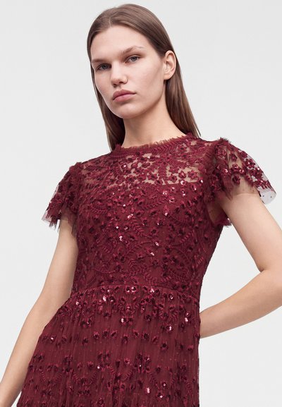 Jeune femme portant une robe en dentelle rouge foncé avec broderies florales et sequins, avec des manches courtes transparentes, debout devant un fond uni.