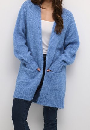 Long, open-front cardigan in light blue soft knit. Features ribbed cuffs and hem, with two front pockets. Worn over a white top.