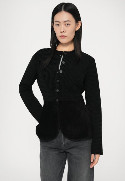 Mlada ženska s hladnimi črnimi lasmi, oblečena v črn gumbast cardigan z teksturo in temno sivo jeans hlače, stoji ob beli ozadju.