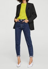 Schwarzes Blazer, leuchtend gelb gestrickter Pullover, hochgeschnittene blaue Jeans mit umgeschlagenen Säumen und weiße spitze Absätze. Minimalistisches Design, lässiger Schnitt.