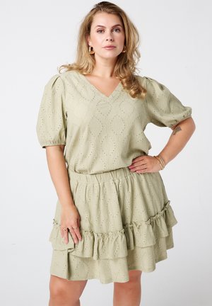 Femme blonde aux cheveux bouclés portant une robe vert clair à broderies anglaises avec manches bouffantes et jupe volantée à étages, debout avec la main sur la hanche.