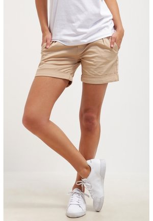 Personne portant un short beige retroussé, un t-shirt blanc et des baskets blanches, debout, jambes croisées et mains dans les poches, sur fond uni.