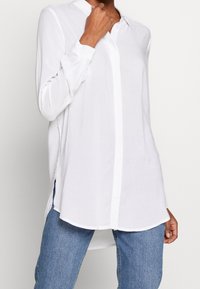 Personne portant un chemisier blanc à manches longues avec des boutons cachés, associé à un jean bleu, la main touchant le col, fond clair uni.