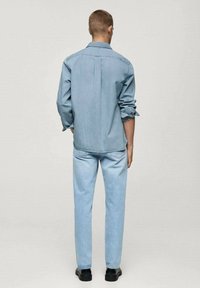 Chemise en denim bleu clair avec col boutonné, manches longues retroussées, associée à un jean bleu clair et des chaussures noires, vue de dos.