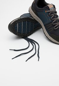 Trailrunning-skor i mörkblått och grått, med en texturerad gummisula, ovandel i mesh, orange detaljer och platta svarta snören.