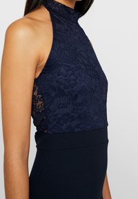 Granatowa sukienka z koronkowym gorsetem o wysokim dekolcie i dopasowaną, gładką spódnicą. Koronka zdobiona jest kwiatowymi wzorami i skomplikowaną fakturą.