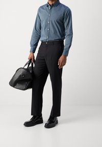 Ciemnoniebieska koszula zapinana na guziki z długimi rękawami, czarne spodnie i wypolerowane czarne buty. Nosi zaokrągloną czarną skórzaną torbę.