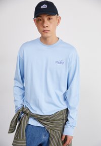 Camisa de manga longa azul claro com o logótipo roxo da "nike", combinada com uma camisa verde e branca com riscas atada à cintura, e um boné escuro.