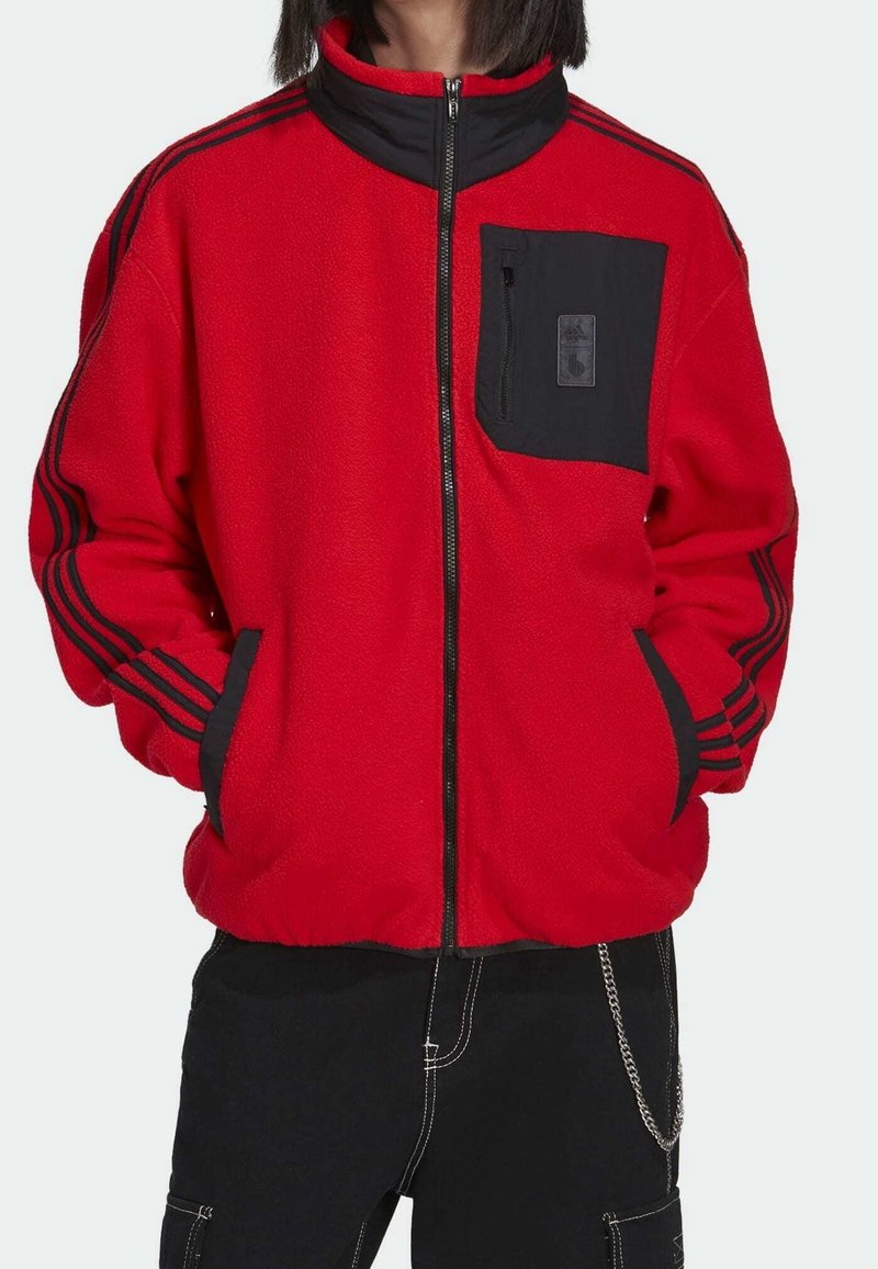 Chaqueta de forro polar roja con detalles negros, cuello alto, bolsillos laterales y un bolsillo en el pecho con un pequeño logo. Tres rayas negras en las mangas.