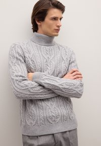 Suéter de punto trenzado gris claro con un cuello alto, que presenta intrincados patrones texturizados y puños y dobladillo acanalados. Llevado con pantalones grises.