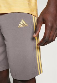 Graue Sportshorts mit gelbem Adidas-Logo und drei gelben Seitenstreifen, ausgestattet mit einer glatten Textur und einem körpernahen Schnitt.