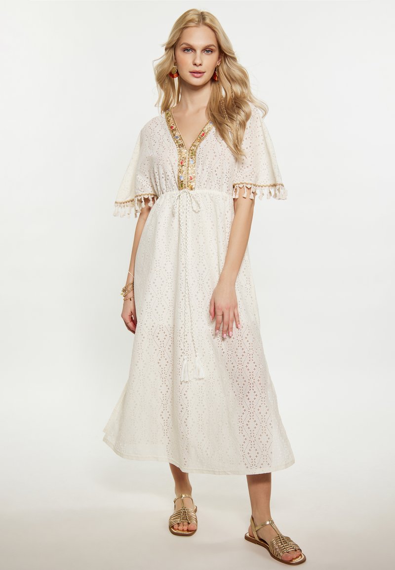 Witte maxi jurk met kantdetails, korte mouwen en een diepe V-hals. Heeft een gouden rand en kwastjesaccenten. Gecombineerd met gouden sandalen.