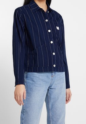 Žena v tmavě modré bundě na knoflíky s bílými vertikálními pruhy a světlých modrých džínách.