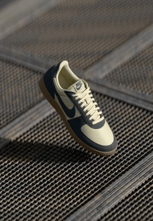 Nike sneaker featuring a cream upper with navy accents, textured rubber sole, and white laces, displayed against a grid surface.