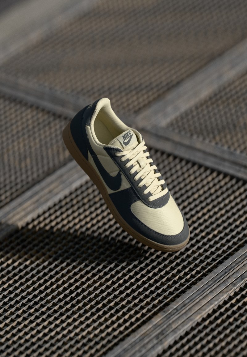 Nike sneaker met een crème bovenkant en marineblauwe accenten, verhoogde rubberen zool en witte veters, weergegeven op een rasteroppervlak.