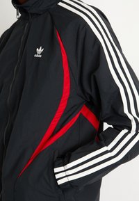 Crni vjetrovka s crvenim i bijelim detaljima, koja ima bijele trostruke trake na rukavima i adidas logo na prsima.