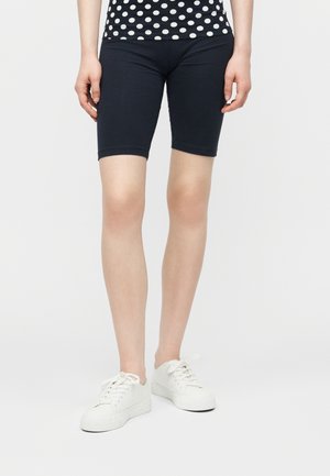 Persona che indossa pantaloncini neri fino al ginocchio, una camicia a pois bianca e nera e scarpe da ginnastica bianche, in piedi davanti a uno sfondo semplice.