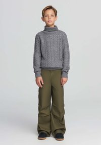 Szary sweter z wysokim kołnierzem w splot warkoczowy, zestawiony z luźnymi oliwkowymi spodniami. Stylizację uzupełniają ściągacze przy rękawach i brązowe sneakersy.
