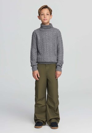 Pullover a collo alto in maglia rasata grigia, abbinato a pantaloni larghi color verde oliva. L'outfit presenta polsini a coste e scarpe da ginnastica marroni.