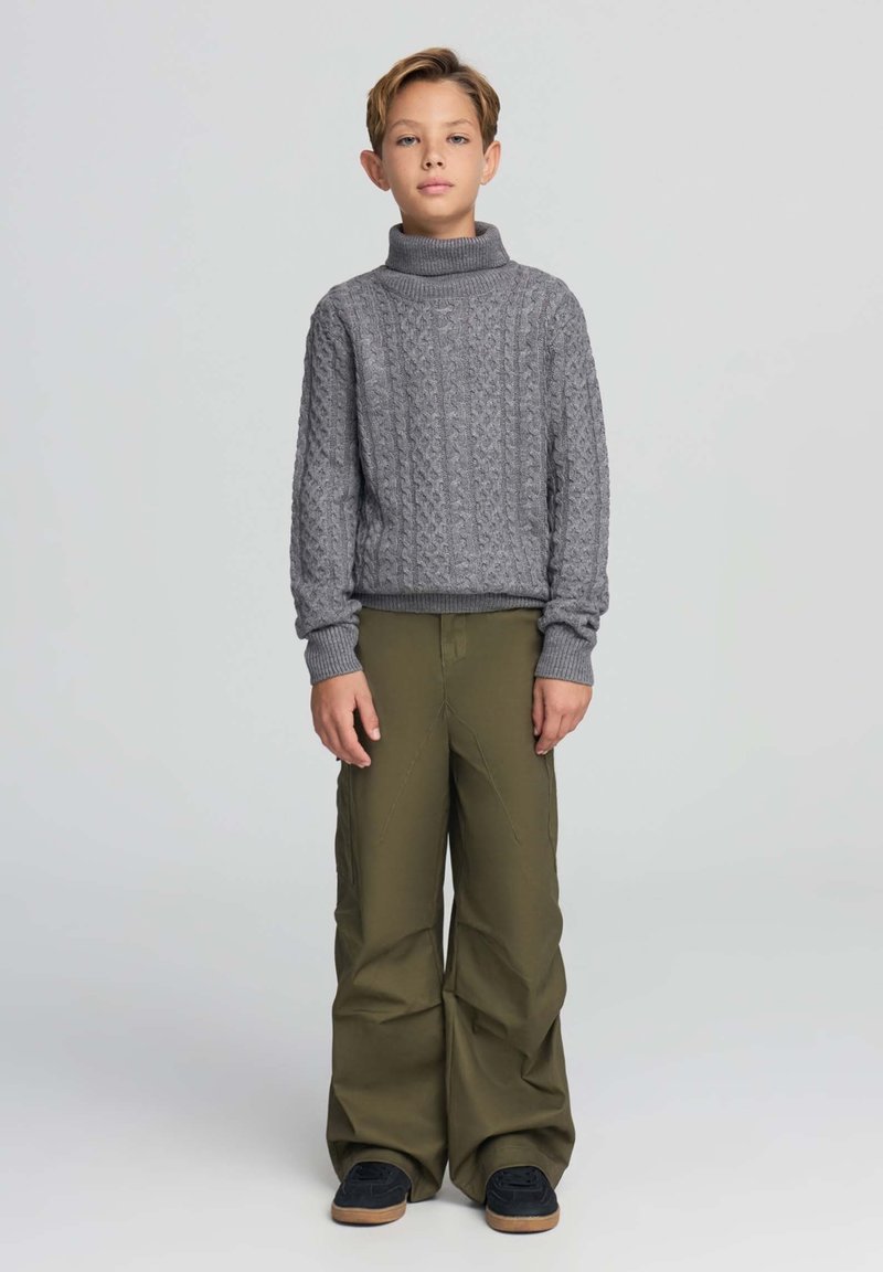 Szary sweter z wysokim kołnierzem w splot warkoczowy, zestawiony z luźnymi oliwkowymi spodniami. Stylizację uzupełniają ściągacze przy rękawach i brązowe sneakersy.