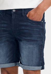 Sötétkék farmer rövidnadrág felhajtott szegéllyel, fém gombos záródással, elöl zsebekkel és enyhén kopott hatású textúrával.