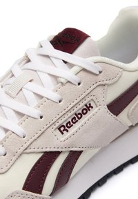 Reebok-Sneaker mit einem Obermaterial aus cremefarbenem Leder und beigefarbenem Wildleder, einem burgunderfarbenen Streifen, weißen Schnürsenkeln und Logodetail an der Seite und Zunge.