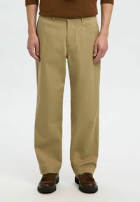 Homme portant un pantalon beige à jambes droites, un pull marron et des mocassins à glands marron foncé sur fond blanc.