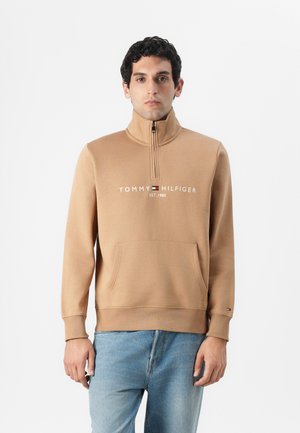 Tamna zip-up dukserica s visokim ovratnikom, prednjim džepom i "Tommy Hilfiger" logom. Ima rebraste rubove na rukavima i donjem dijelu. Nosena preko plavih traperica.