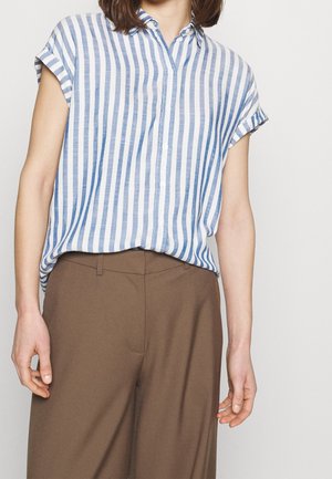 Kobieta ubrane w białą koszulę z krótkim rękawem i pionowymi niebieskimi paskami, wpuszczoną do spodni o wysokim stanie, brązowych, szerokich nogawkach.