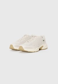 Lacoste sneakers in light beige, featuring a wave texture on the upper, lace-up design, and a cream rubber sole with a green logo accent.