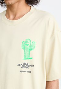 Beige bomull T-skjorte med et grønt kaktusgrafikk og svart tekst "ALL CONDITIONS GEAR Big Bend, TEXAS" på forsiden. Rund hals.