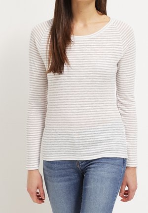 Femme portant une chemise rayée blanche et gris clair à manches longues et un jean bleu, visible du cou jusqu'aux hanches sur un fond uni.