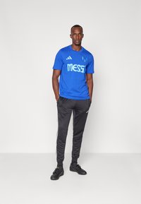 Blaues T-Shirt mit kurzen Ärmeln und dem weißen Text "MESSI", kombiniert mit schwarzen Jogginghosen mit weißen Seitestreifen und schwarzen Sportschuhen.