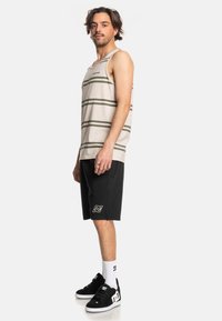 Beige tank top with green stripes, black shorts, and black sneakers. Features a logo on the shorts, worn with white socks.