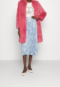 Vrouw in een felroze nepbontjas, witte t-shirt met logo, blauwe geplooide rok met grote bloemenprint en witte slingback hakken.