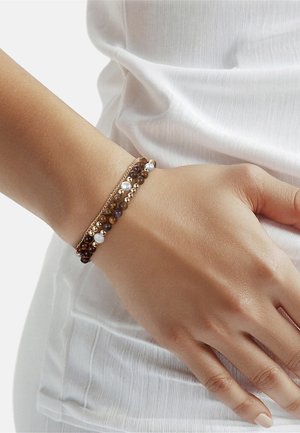 Pols met drie armbanden: bruin en goudkleurige kralen gemengd met kleine witte parels, afgebeeld tegen een wit textuurshirt en een lichte achtergrond.