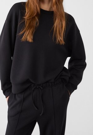 Femme portant un sweat-shirt noir à manches longues et un pantalon de jogging noir à cordon, les mains dans les poches, sur un fond clair et uni.