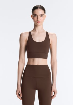 Mladá žena so zviazanými vlasmi, oblečená v hnedom športovom podprsenke a legínach s vysokým pásom, stojaca na jednotnom svetlom pozadí.