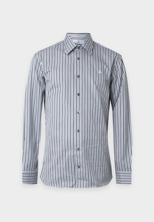 Ung mand stående iført en sort- og hvidstribet skjorte med knapper, beige bukser med opbuk og sorte sko på hvid baggrund.
