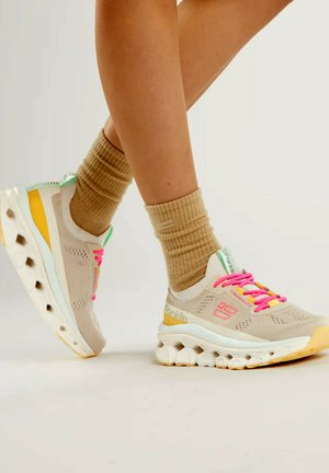 Sneakers beige con parte superiore in rete, lacci rosa neon, dettagli verdi e una suola bianca chunky con ritagli circolari e un tallone giallo.