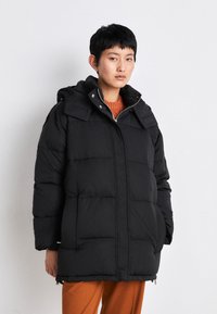 Svart puff jakke med en firkantet fasong og hette. Har horisontal quilting, glidelåslukking og to sidelommer. Brukt over en oransje genser.
