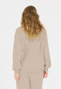 Beige collegepaita, joka on rentoa mittaa, resorihihnoilla ja helmalla. Kangas on sileä, esitetty takaa, jossa näkyy laineikas hius.