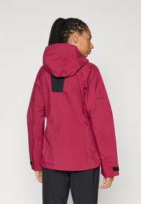 GORE-TEX jakke i dyb rød med et sort panel på ryggen. Har en hætte, tekstureret stof og justerbare manchetter. Letvægt og funktionel.