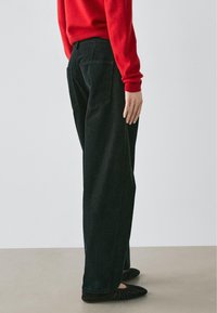 Jean noir à jambes larges avec une finition texturée et deux poches arrière, associé à un pull rouge et des ballerines noires, vu de profil.