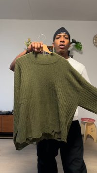 Olivengrøn strikket sweater med ribstrikket tekstur, oversized pasform, høj hals og frynset kant, holdt på en bøjle af en person.