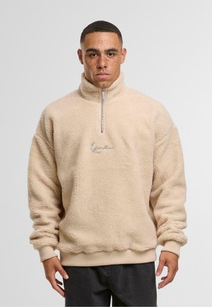 Beiger Fleece-Pullover mit hohem Kragen und halbseitigem Reißverschluss. Mit gerippten Bündchen und Saum sowie einem gestickten Logo auf der Brust.
