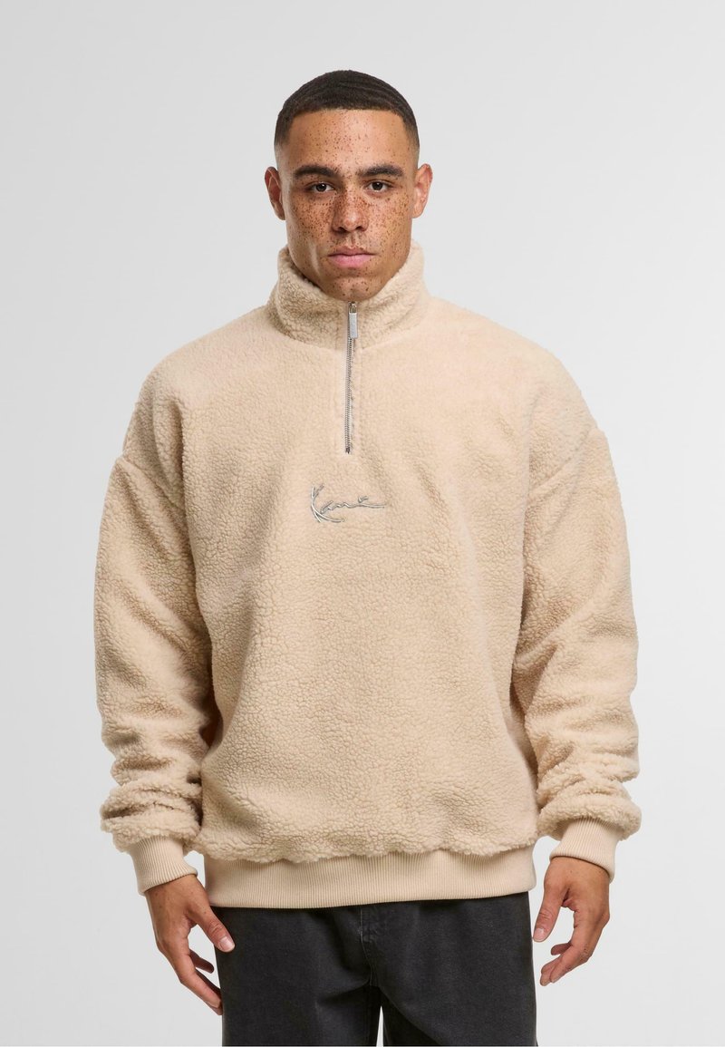 Pulover din fleece bej cu guler înalt și închidere cu fermoar pe jumătate. Prezintă manșete și tiv ribbate, precum și un logo brodat pe piept.
