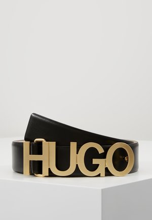 Schwarzer Ledergürtel mit großer goldener Schnalle mit der Aufschrift „HUGO“ auf weißer Oberfläche präsentiert.