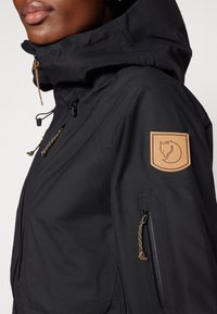 Veste noire résistante aux intempéries avec un col haut, une capuche ajustable et une pièce en cuir beige sur l'épaule. Comprend des poches zippées.